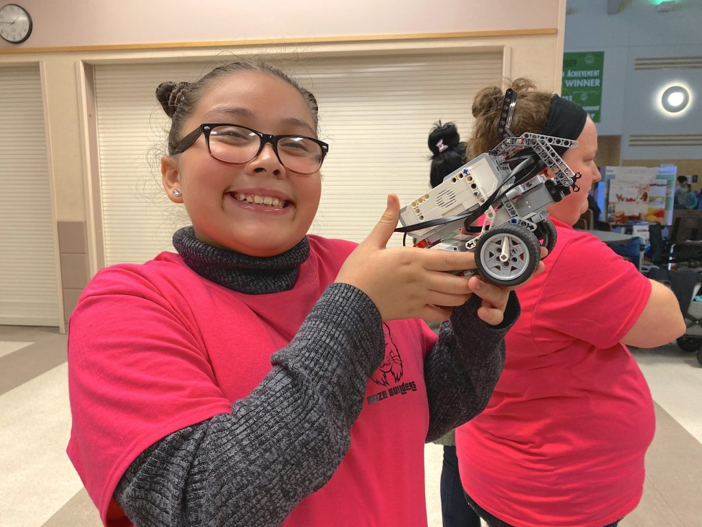 Young, smiling female student in bright pink shirt holds up a robot on wheels